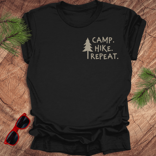 A black t-shirt with the text 'CAMP. HIKE. REPEAT with a pine tree next to it. the shirt is on a wood background with pine leaves and a pair of red sunglasses.