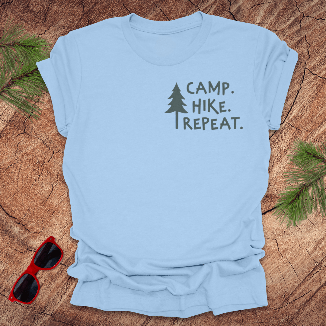 A baby blue t-shirt with the text 'CAMP. HIKE. REPEAT with a pine tree next to it. the shirt is on a wood background with pine leaves and a pair of red sunglasses.