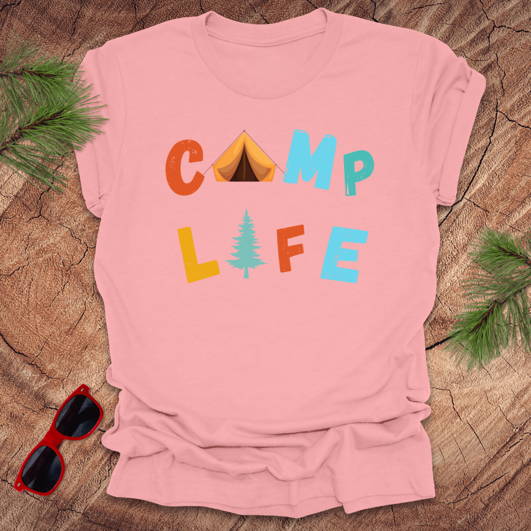 pink t-shirt with 'Camp Life' text graphic, featuring colorful letters and a pine tree, displayed on a wooden background with pine needles and red sunglasses.