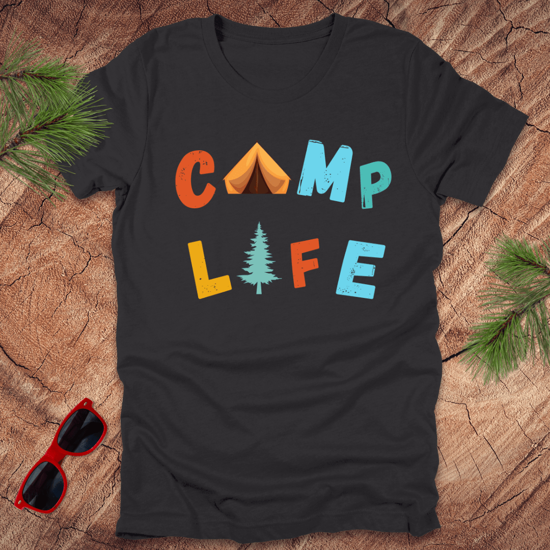 Black t-shirt with 'Camp Life' text graphic, featuring colorful letters and a pine tree, displayed on a wooden background with pine needles and red sunglasses.