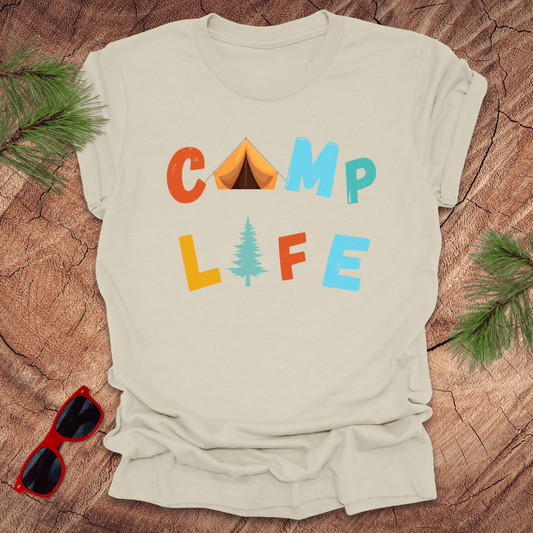 natural t-shirt with 'Camp Life' text graphic, featuring colorful letters and a pine tree, displayed on a wooden background with pine needles and red sunglasses.