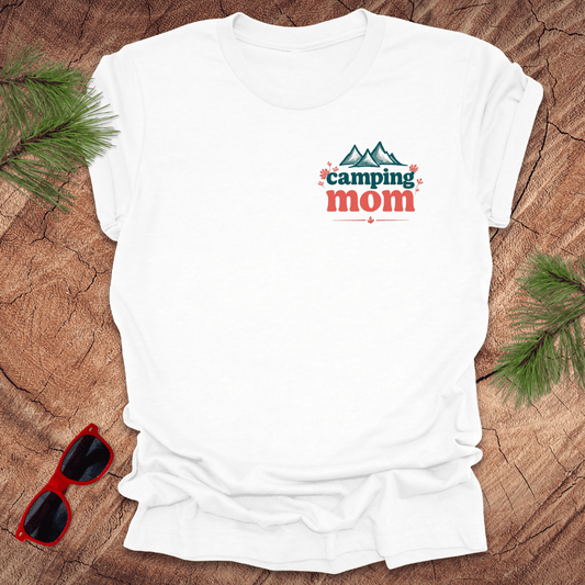 A white t-shirt with 'Camping Mom' text and a minimalist mountain design in blue on the front, displayed on a wooden surface with pine needles and red sunglasses to the side.
