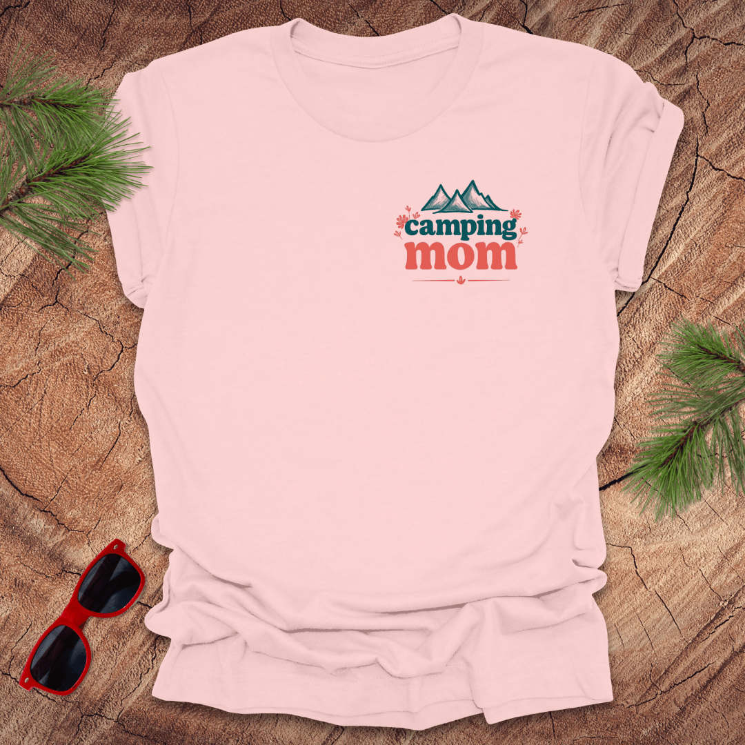 A pink t-shirt with 'Camping Mom' text and a minimalist mountain design in blue on the front, displayed on a wooden surface with pine needles and red sunglasses to the side.