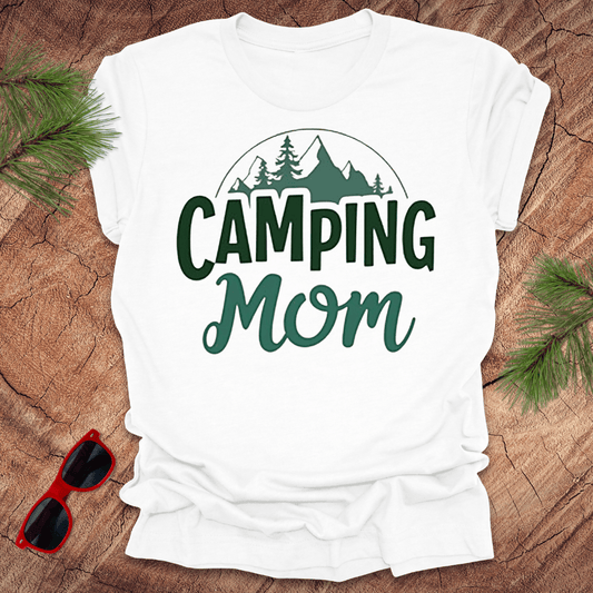 A white t-shirt with 'CAMPING MOM' printed in green and mountain silhouettes in a minimalist style. The shirt is presented on a wooden surface with pine needles around it and a pair of sunglasses to the side.
