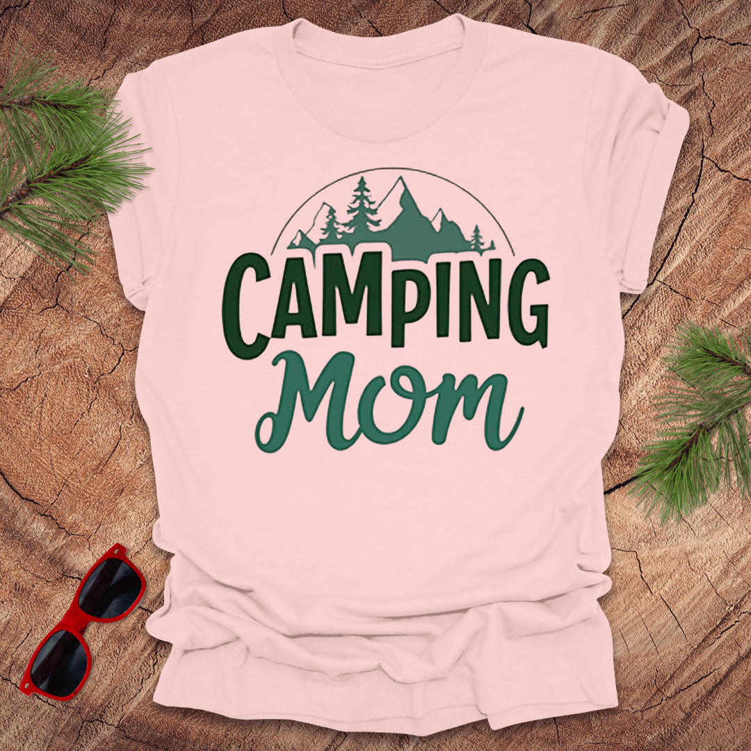 A  pink t-shirt with 'CAMPING MOM' printed in green and mountain silhouettes in a minimalist style. The shirt is presented on a wooden surface with pine needles around it and a pair of sunglasses to the side.