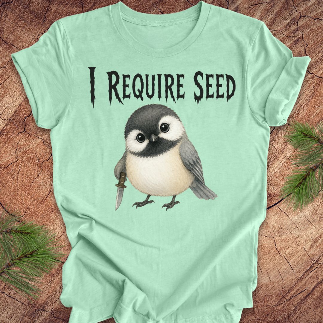 An mint green short-sleeve T-shirt laid flat on a wood surface with autumn leaves around it. The shirt has black text reading 'I REQUIRE SEED' above a cartoon chickadee holding a dagger.