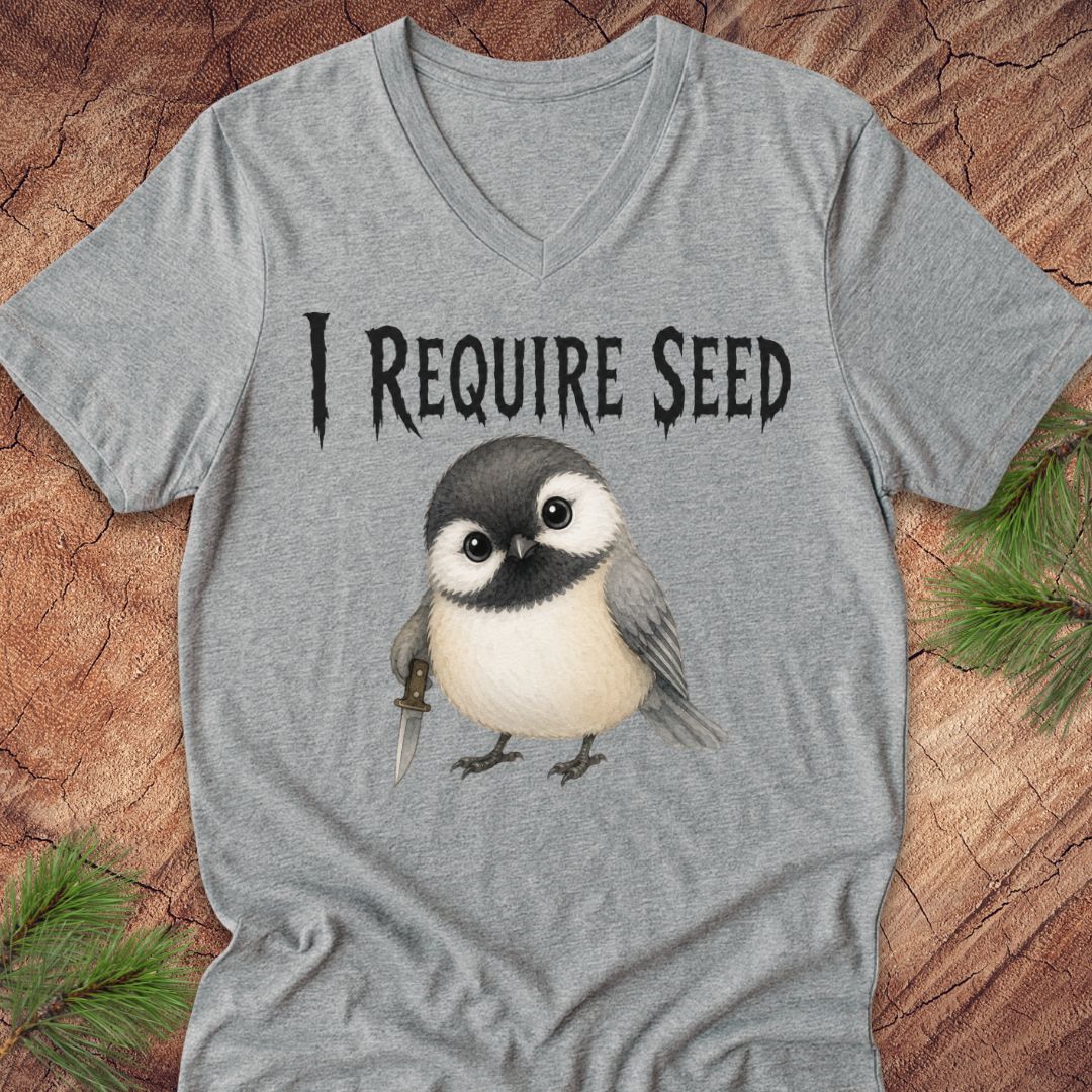 A heather gray V-neck t-shirt laid flat on a rustic wood background with pine branches in the corners. The shirt features a graphic of a cute chickadee bird holding a seed, with the text 'I REQUIRE SEED' printed above it.