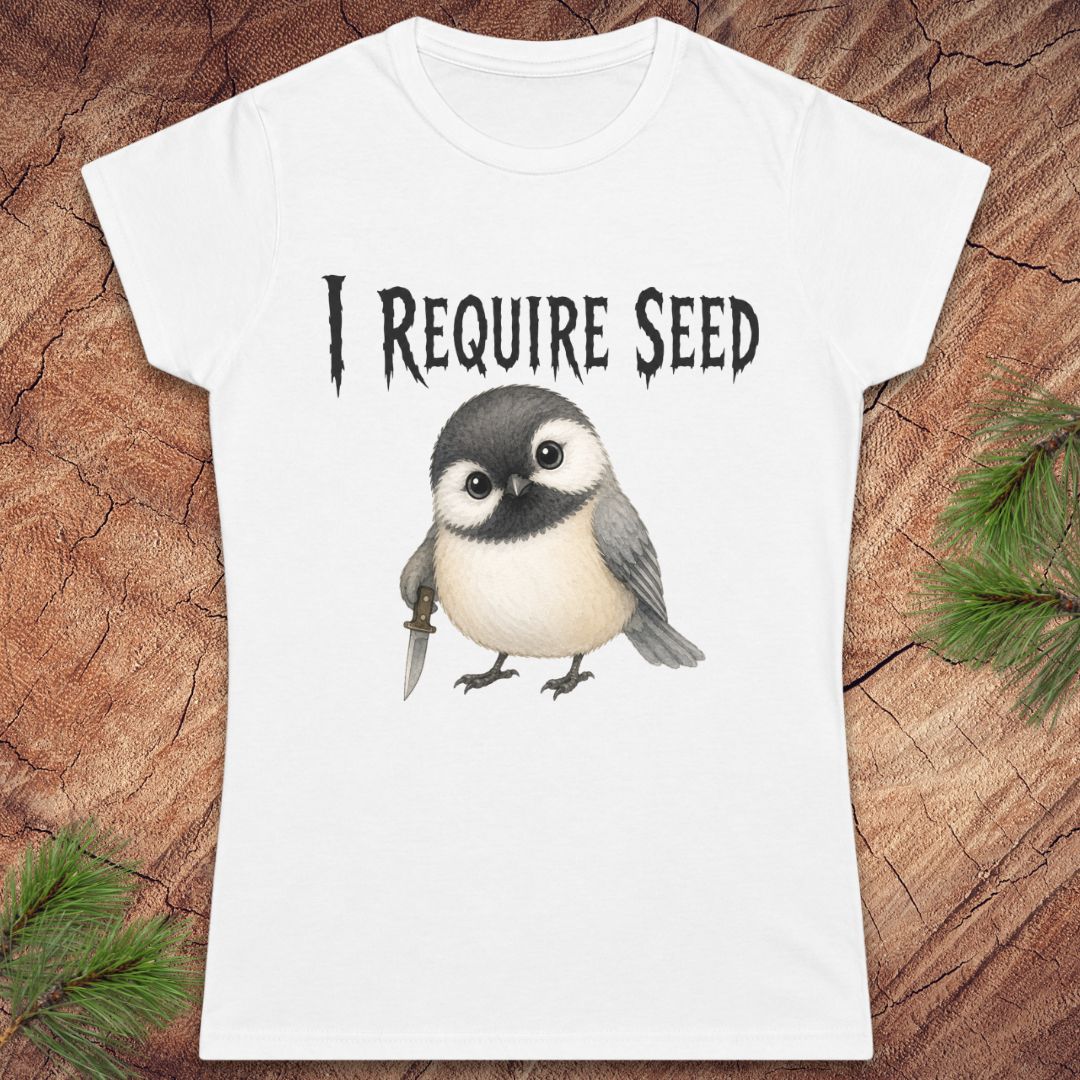 A white short-sleeve t-shirt with a graphic of a cute chickadee holding a dagger and the text 'I REQUIRE SEED' in a gothic font, displayed flat on a rustic wood background with pine needles.