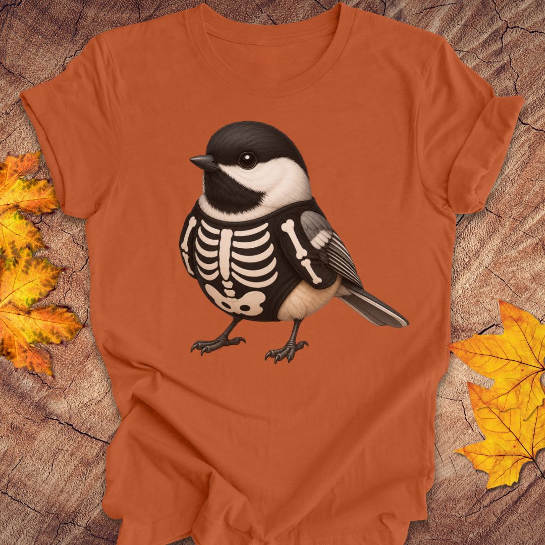 An orange short-sleeve t-shirt laid flat on a rustic wood background with autumn leaves. The shirt features a graphic of a black and white chickadee bird with a skeleton print on its chest.