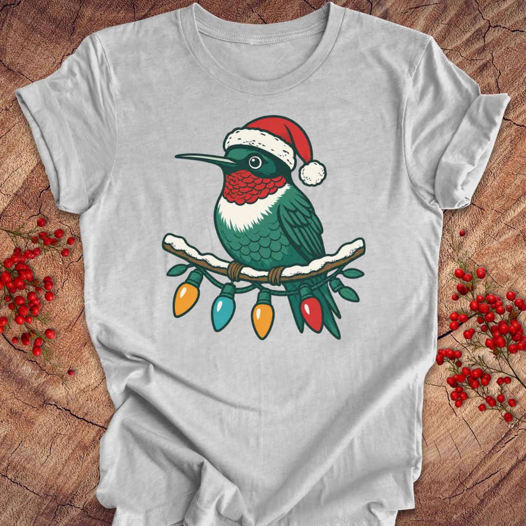 A light gray short-sleeve t-shirt laid flat on a rustic wood background, featuring a graphic of a green hummingbird wearing a red Santa hat perched on a branch with multicolored Christmas lights.