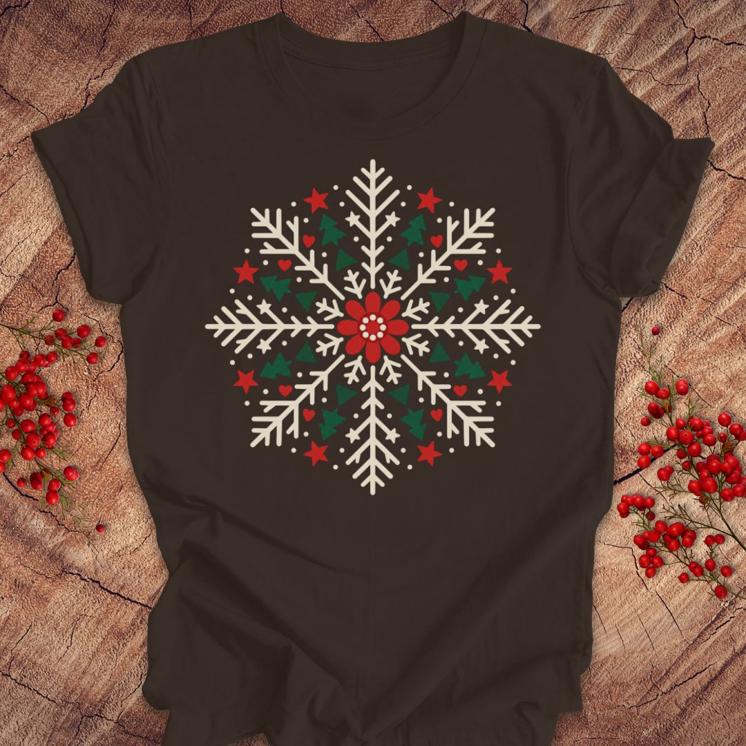 Brown t-shirt with a creamy white folk-art snowflake filled with tiny green trees, red stars, and hearts around a red flower, laid flat on a rustic wood background with red winter berries.