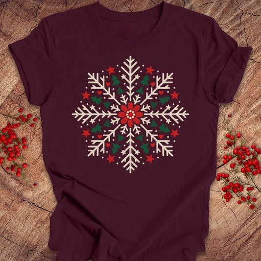 Maroon t-shirt with a creamy white folk-art snowflake filled with tiny green trees, red stars, and hearts around a red flower, laid flat on a rustic wood background with red winter berries.
