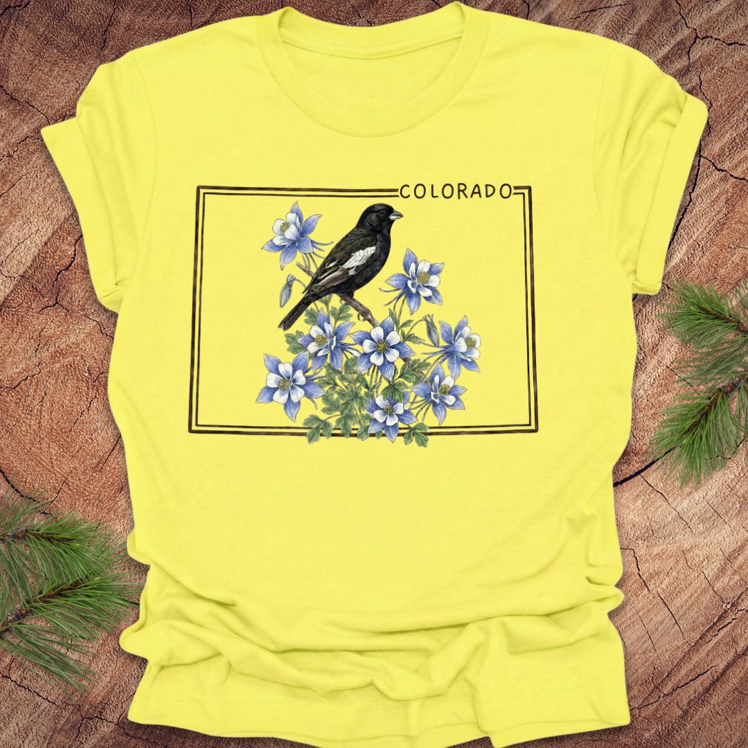 Yellow t-shirt with the outline of Colorado and lark bunting bird and blue flowers graphic on a wooden surface