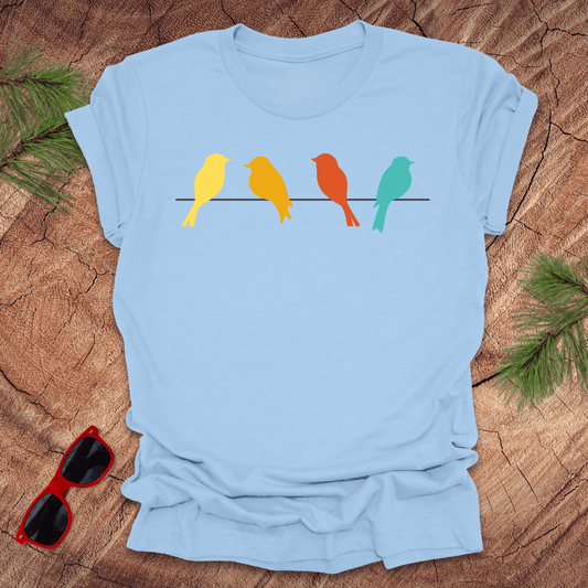 A graphic blue t-shirt with a unisex fit featuring vibrant bird silhouettes in yellow, orange, and blue colors on a beige background, displayed on a wooden surface with green foliage and red sunglasses in the background.