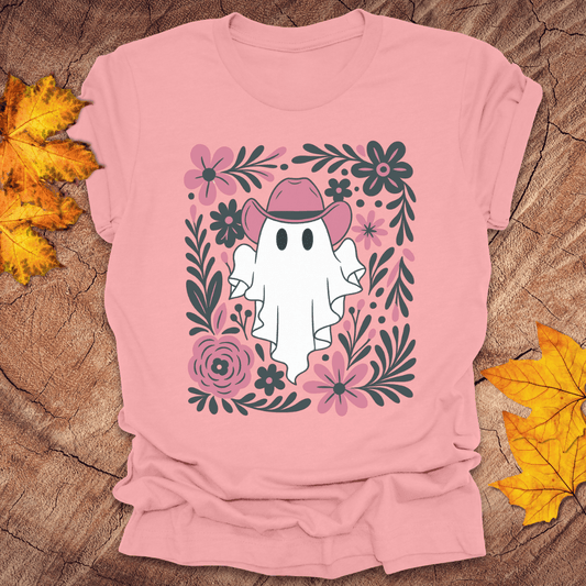 Pink t-shirt with a ghost wearing a cowboy hat and surrounded by flowers on a wooden surface with fall leaves.