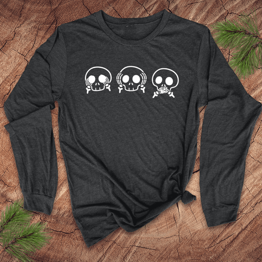 Dark gray long-sleeve tshirt with 3 skulls simulating see no evil, hear no evil, speak no evil, on a wood background with pine leaves.