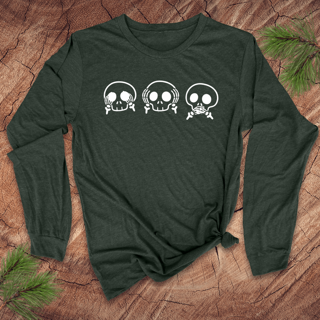Green long-sleeve tshirt with 3 skulls simulating see no evil, hear no evil, speak no evil, on a wood background with pine leaves.