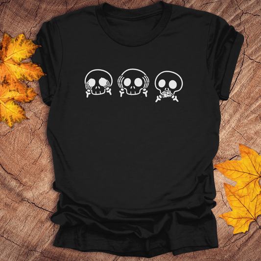 Black tshirt with 3 skulls simulating see no evil, hear no evil, speak no evil, on a wood background with fall leaves.