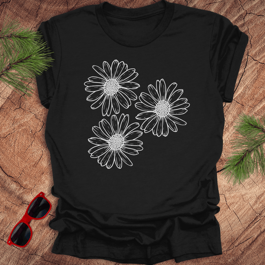A black t-shirt with 3 daisies prominently displayed. on a wooden surface with sunglasses and greenery.