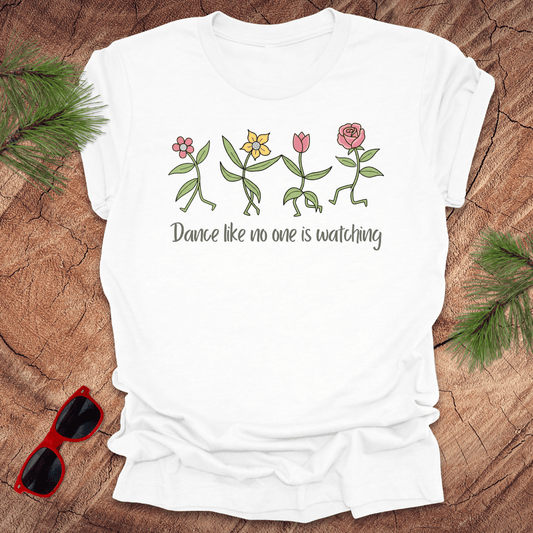 White t-shirt with flower dancing design and text, 'Dance like no one is watching' on a wooden surface with sunglasses and greenery.