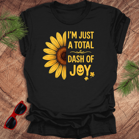 Black t-shirt with sunflower graphic and text 'i'm just a total dash of joy' on a wooden surface with sunglasses and pine branches.