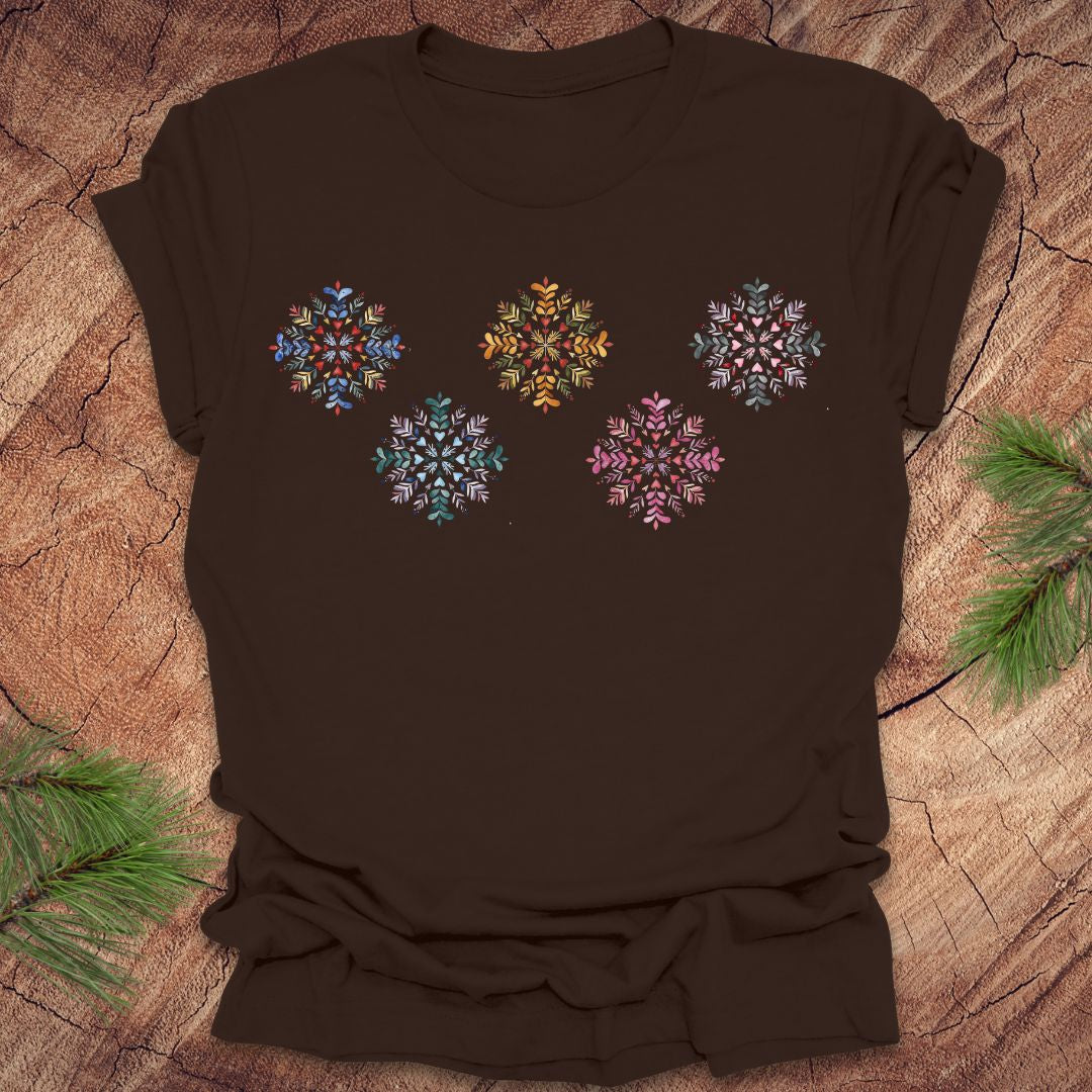 Brown t-shirt with colorful folk art snowflake designs on a wooden surface with pine branches.