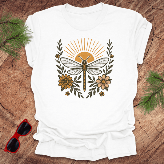 White t-shirt with a dragonfly and floral design on a wooden surface with sunglasses and greenery.