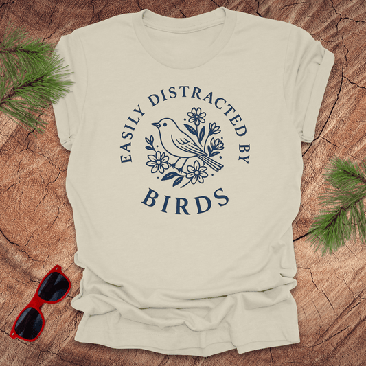 Beige t-shirt with 'Easily Distracted by Birds' design on a wooden surface with sunglasses and greenery.