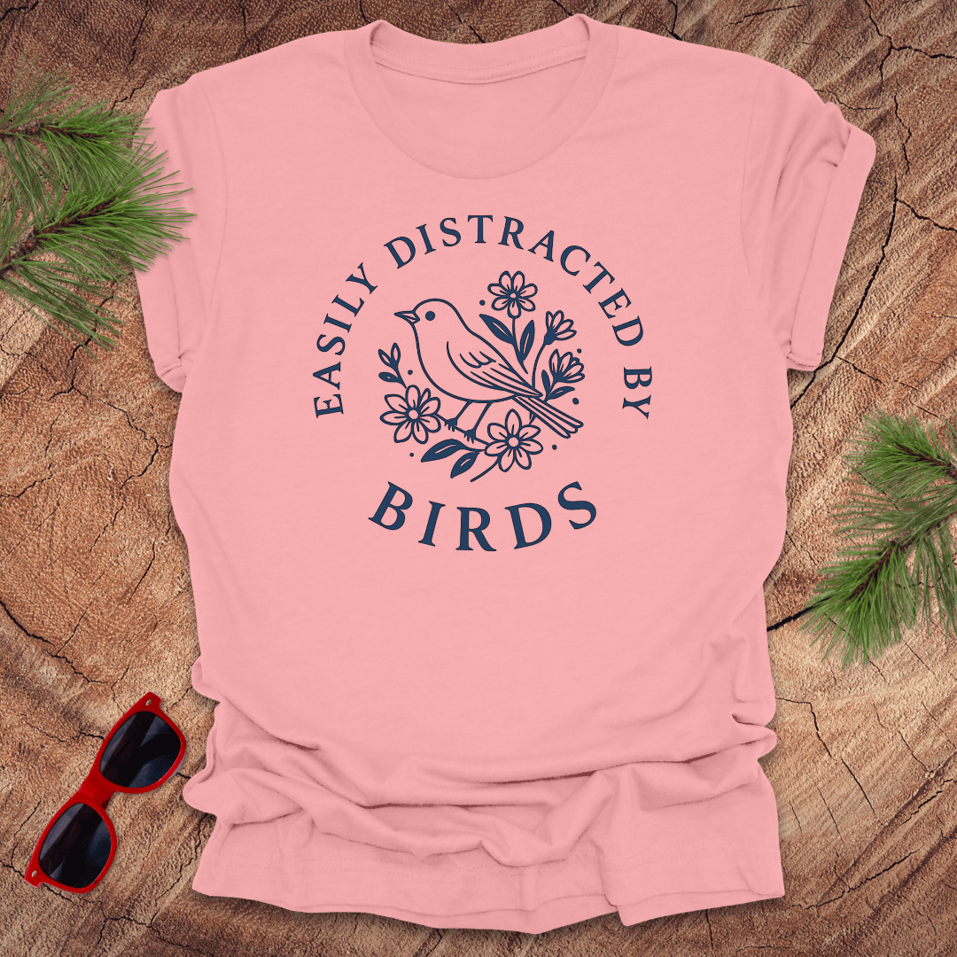 Pink t-shirt with 'Easily Distracted by Birds' design on a wooden surface with sunglasses and greenery.