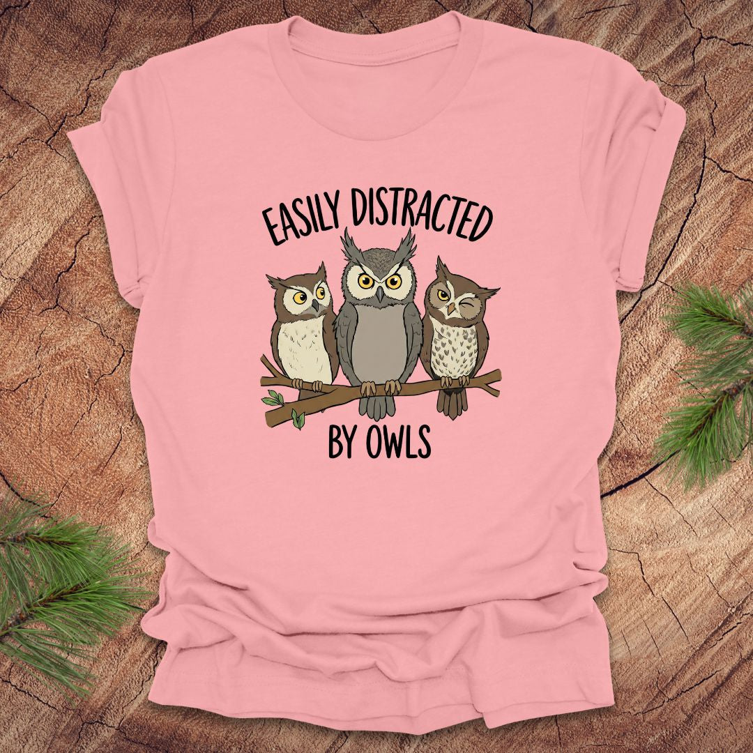 Pink t-shirt with owl design and text 'Easily Distracted by Owls' on a wooden surface.