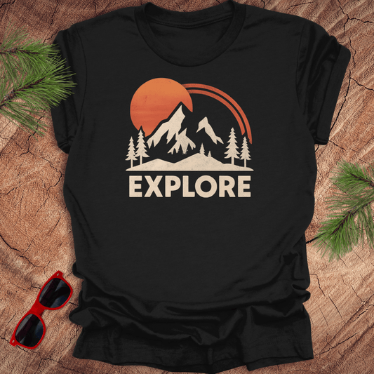 Black t-shirt with mountain and sun design and 'EXPLORE' text on a wooden surface with sunglasses and pine branches.