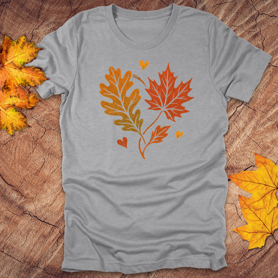 Gray t-shirt with autumn leaf design in the shape of a heart on a textured surface with fall leaves.