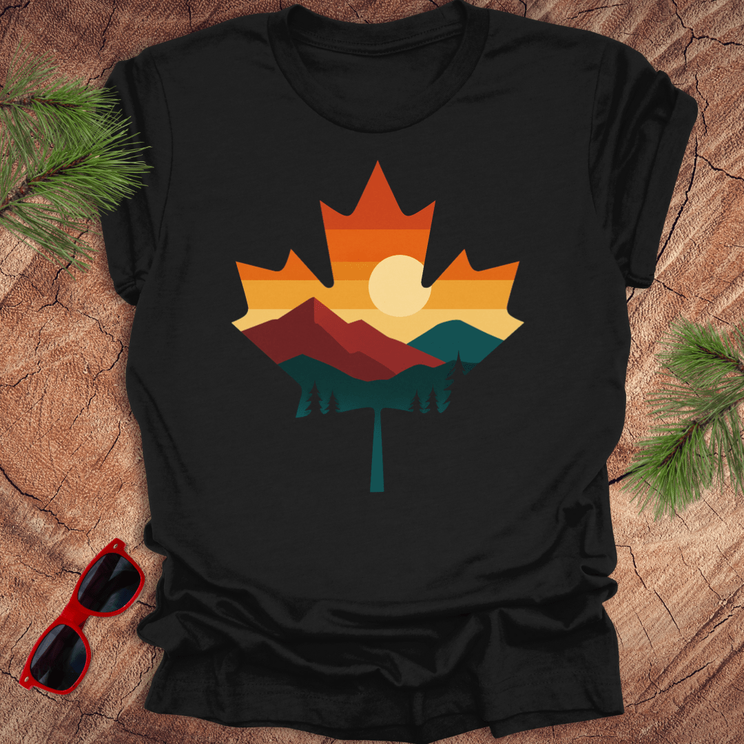 Black t-shirt with a colorful maple leaf design featuring mountains and a sun, placed on a wooden surface with sunglasses and pine branches.