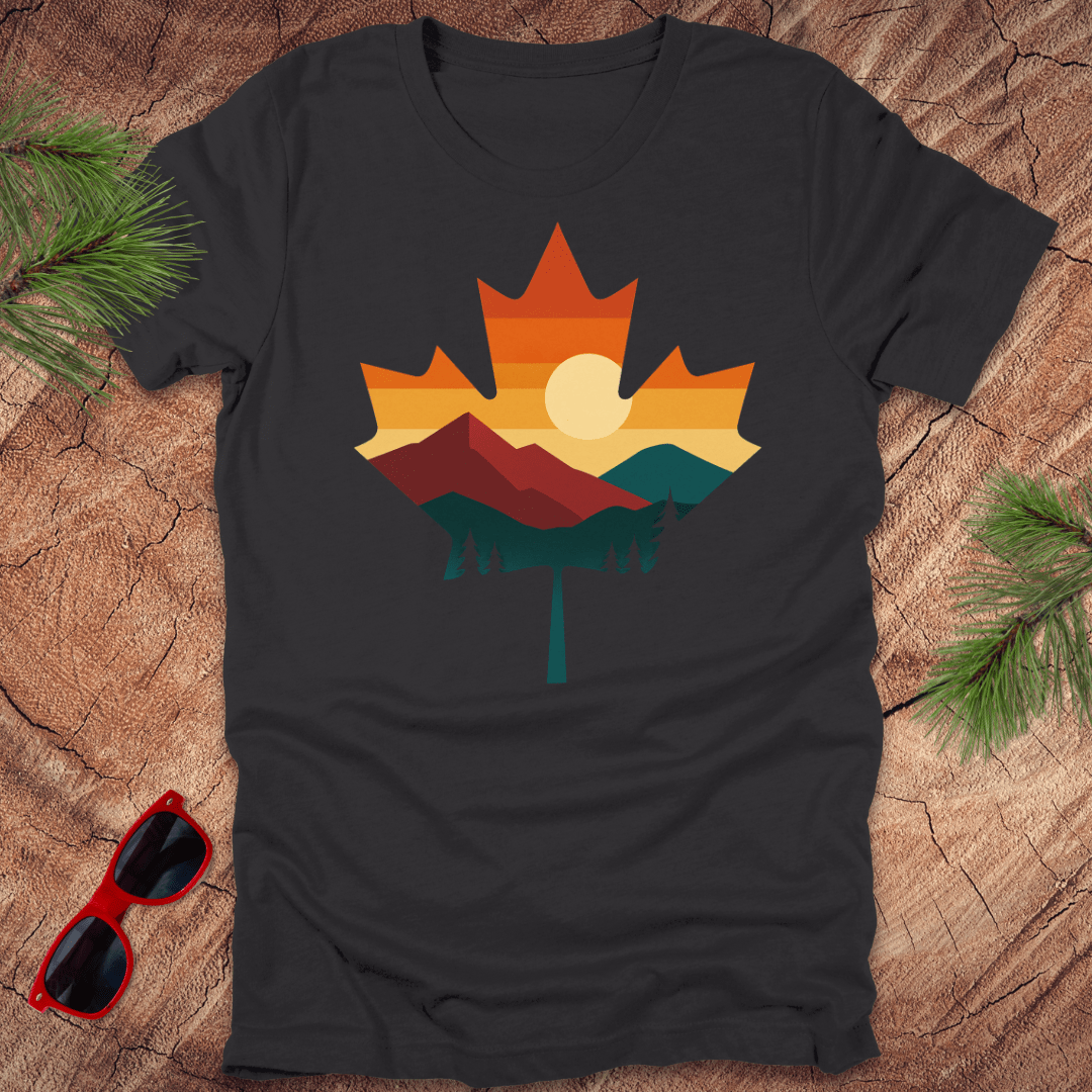 Dark gray t-shirt with a colorful maple leaf design on a wooden surface with sunglasses and pine branches.