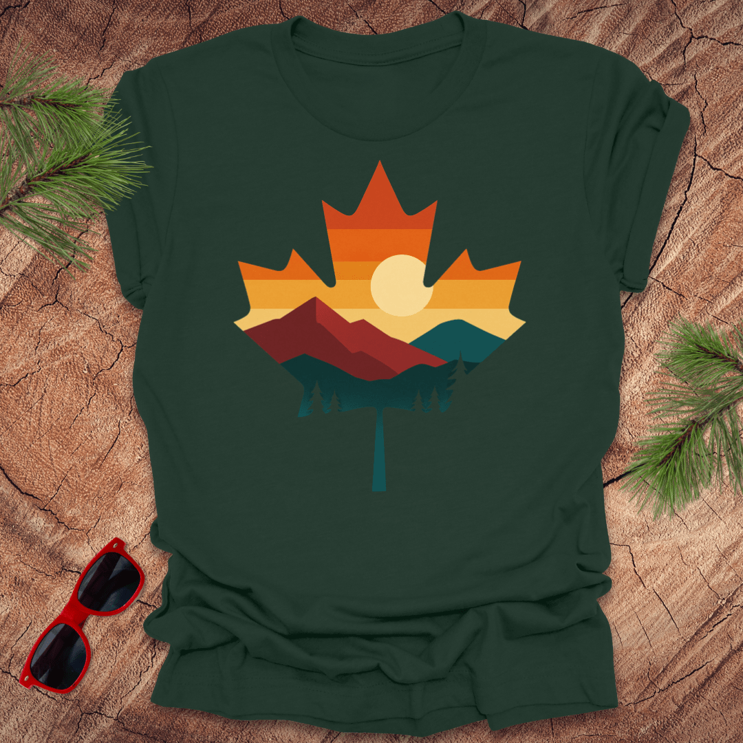 Green t-shirt with a colorful maple leaf design on a wooden surface with sunglasses and pine branches.