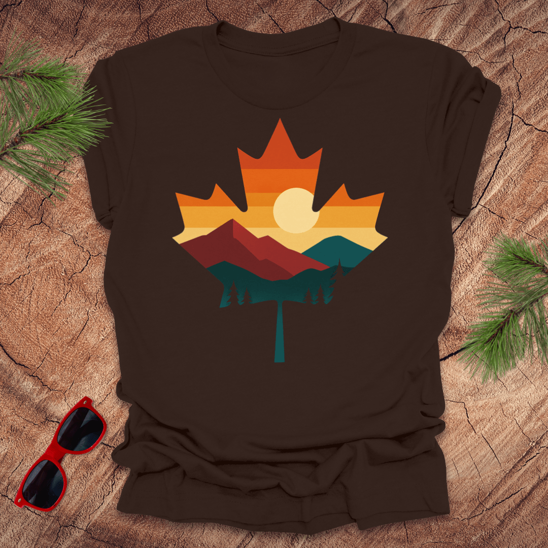 Brown t-shirt with a colorful maple leaf design on a wooden surface with sunglasses and pine branches.