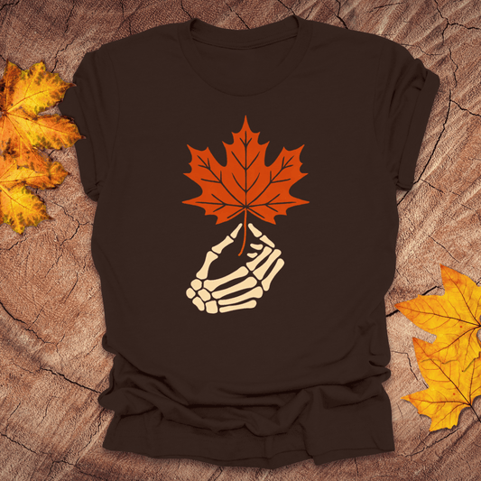 Brown t-shirt with skeleton hand holding an orange maple leaf on a textured brown background with autumn leaves.