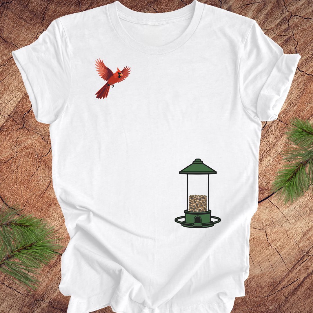A white short-sleeve crewneck t-shirt laid flat on a rustic wood background, featuring a graphic of a red cardinal bird and a green bird feeder. Pine needles are visible in the corners.