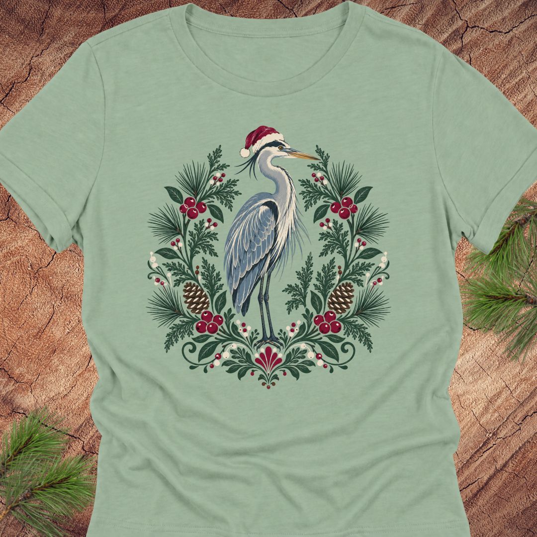 A sage green short-sleeve t-shirt featuring a festive graphic of a blue heron wearing a red Santa hat, surrounded by a wreath of green pine needles, red berries, and pinecones, laid flat on a rustic wood background with pine branches in the corners.