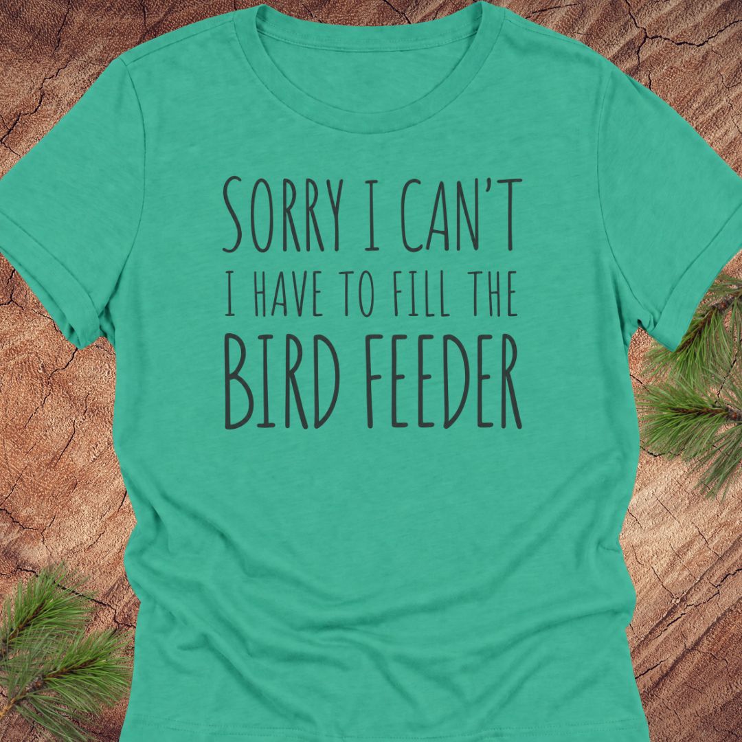 A teal green short-sleeve t-shirt with the text 'SORRY I CAN'T I HAVE TO FILL THE BIRD FEEDER' printed in black on the front, laid flat on a rustic wood background with pine needles in the corners.