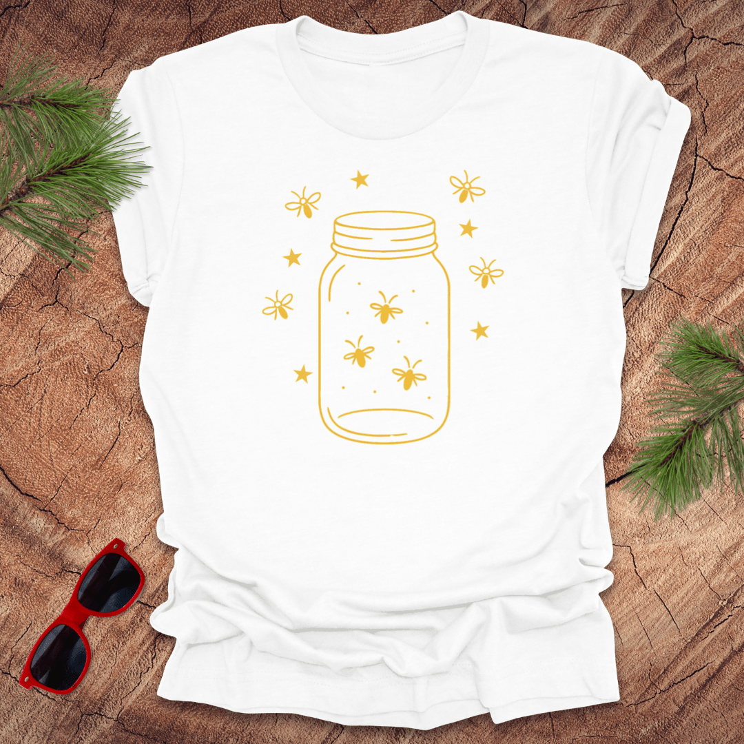 White t-shirt with a yellow jar and firefly design on a wooden surface with sunglasses and greenery.