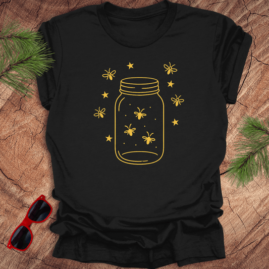 Black t-shirt with a yellow jar and firefly design on a wooden surface with sunglasses and greenery.