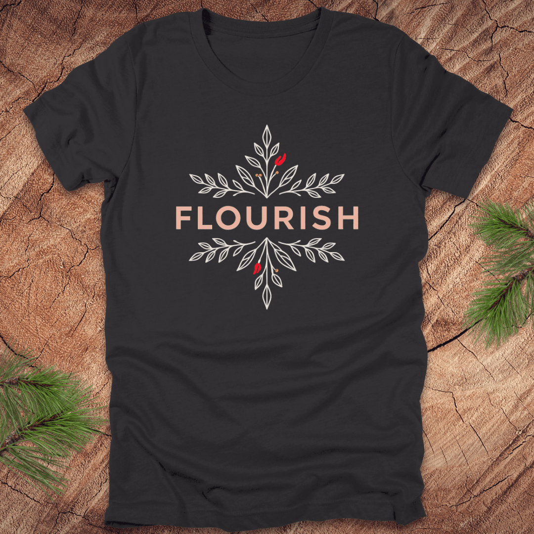 Dark grey t-shirt with 'FLOURISH' design and flowers on a wooden surface with greenery.