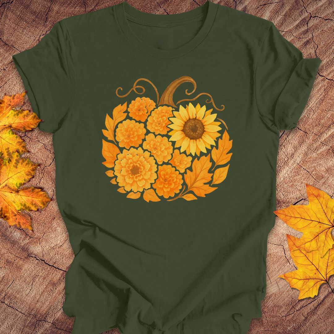 A dark olive green short-sleeve t-shirt with a graphic of a pumpkin made of yellow sunflowers and orange marigolds, styled flat on a rustic wood background with autumn leaves.