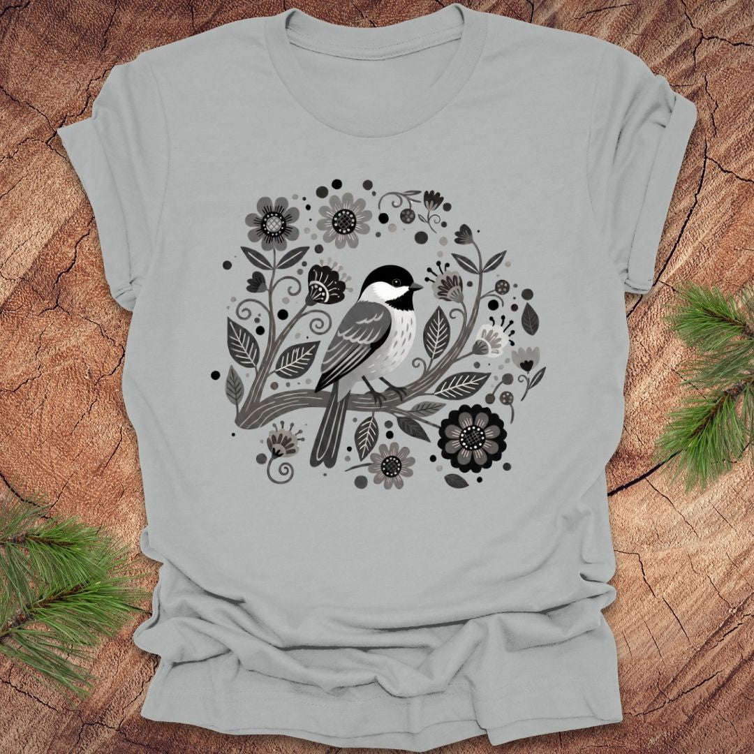 Silver t-shirt of a chickadee on a branch surrounded by folk art flowers, all in black and white, on a wooden background.
