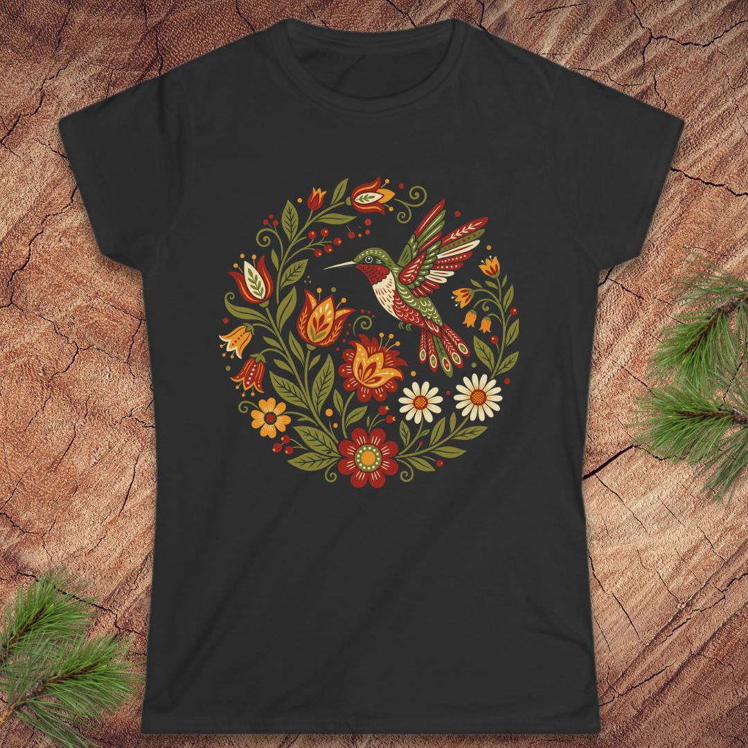 A black short-sleeve crewneck t-shirt featuring a colorful circular graphic of a hummingbird surrounded by flowers and leaves, laid flat on a rustic wood background with pine needles in the corners.