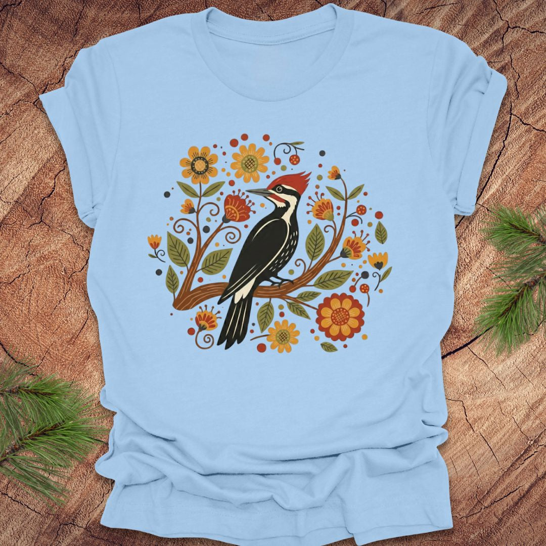Baby blue T-Shirt with a pileated woodpecker on a branch surrounded by folk art flowers on a wood background.