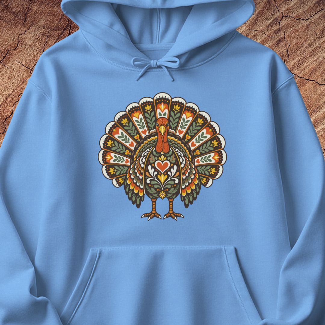 A light blue pullover hoodie laid flat on a rustic wood background, featuring a large decorative turkey graphic on the chest in orange, green, beige, and brown tones.