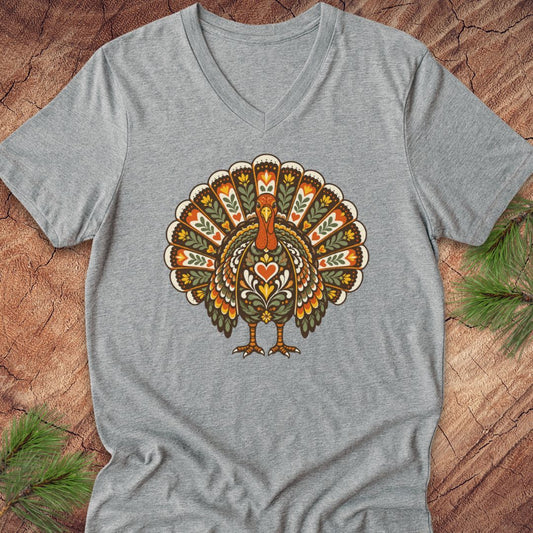 A heather gray V-neck t-shirt featuring a colorful, patterned graphic of a turkey on the chest, laid flat on a rustic wood background with pine needles.