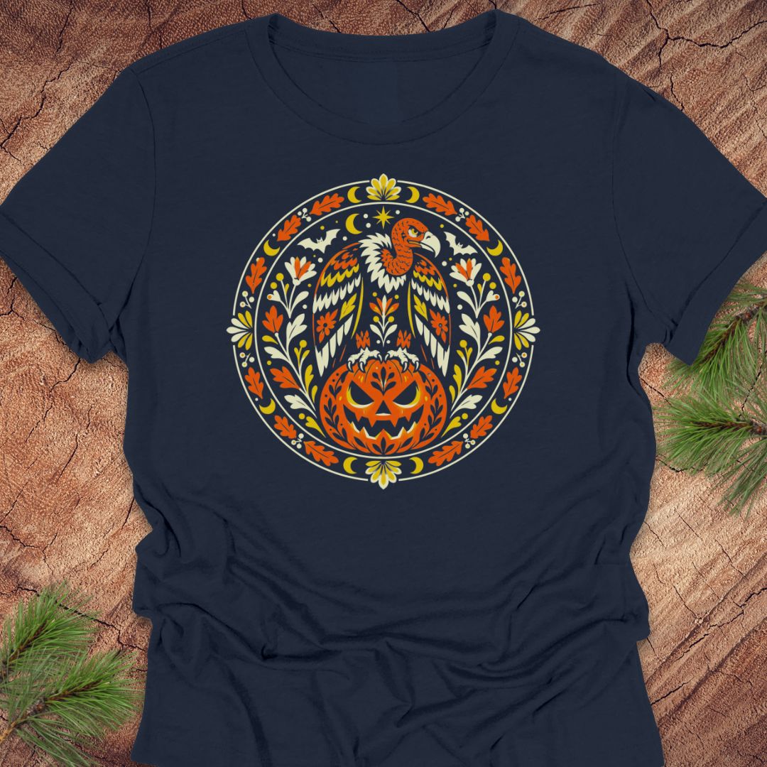 A navy blue short-sleeve t-shirt with a circular graphic of a bald eagle perched on a carved jack-o'-lantern pumpkin, surrounded by autumn leaves and a crescent moon, laid flat on a rustic wood background with pine branches.
